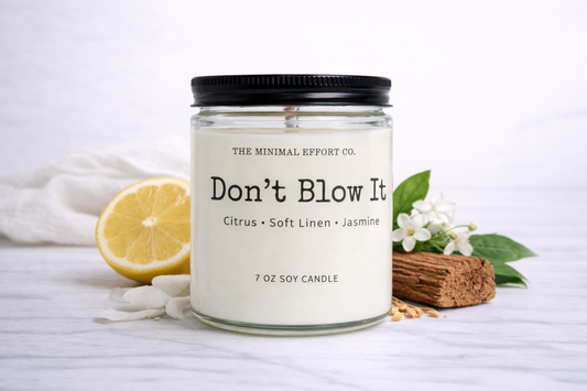 Candle jar labeled 'Don't Blow It' with lemon, flowers, and wood on a white background