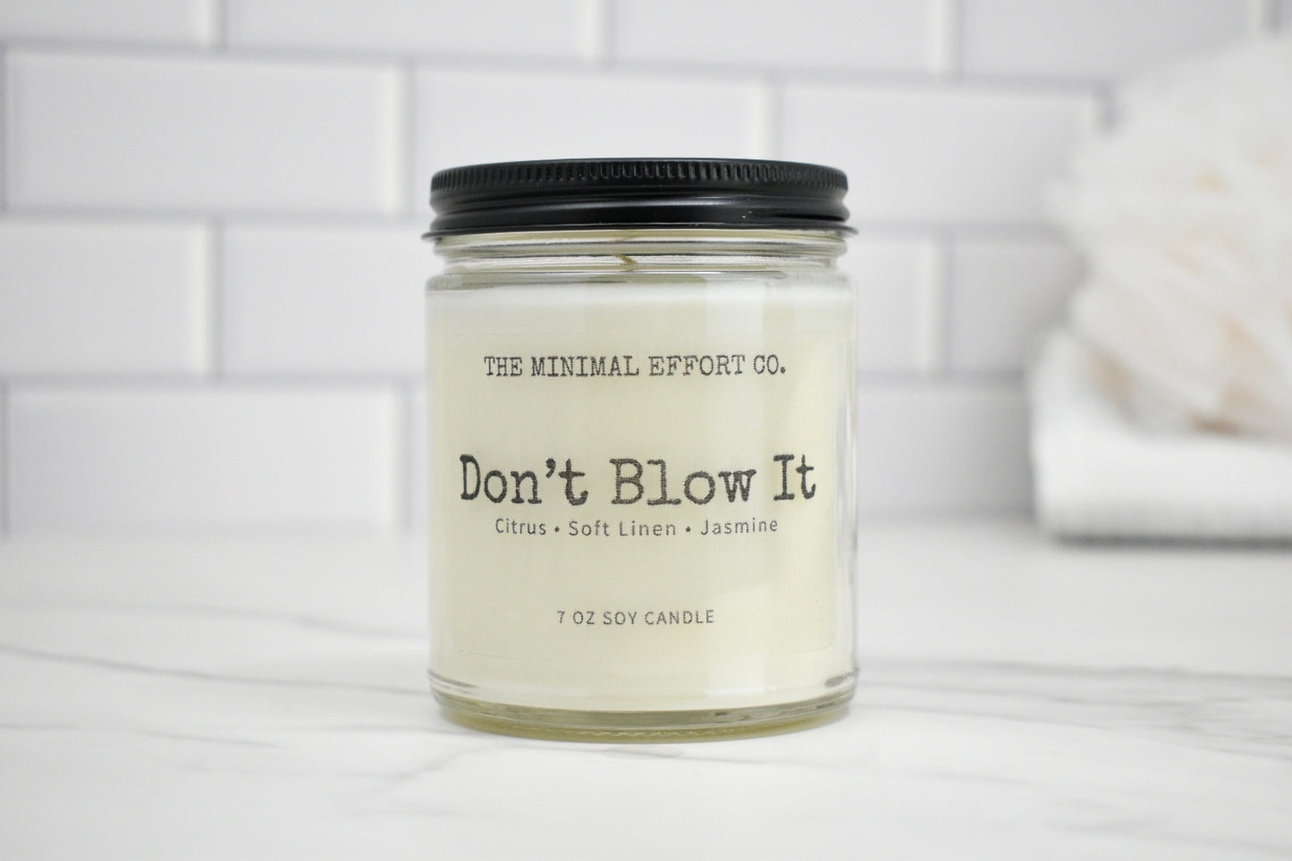 Candle jar labeled 'Don't Blow It' by The Minimal Effort Co. on a white surface with a tiled wall background.