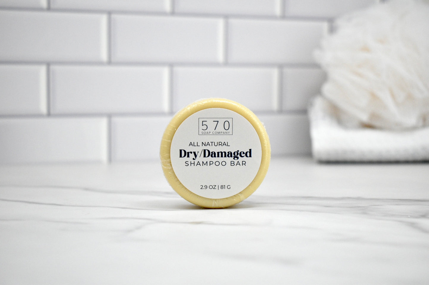 Shampoo bar labeled '570 All Natural Dry Damaged' on a white surface with a tiled wall background.