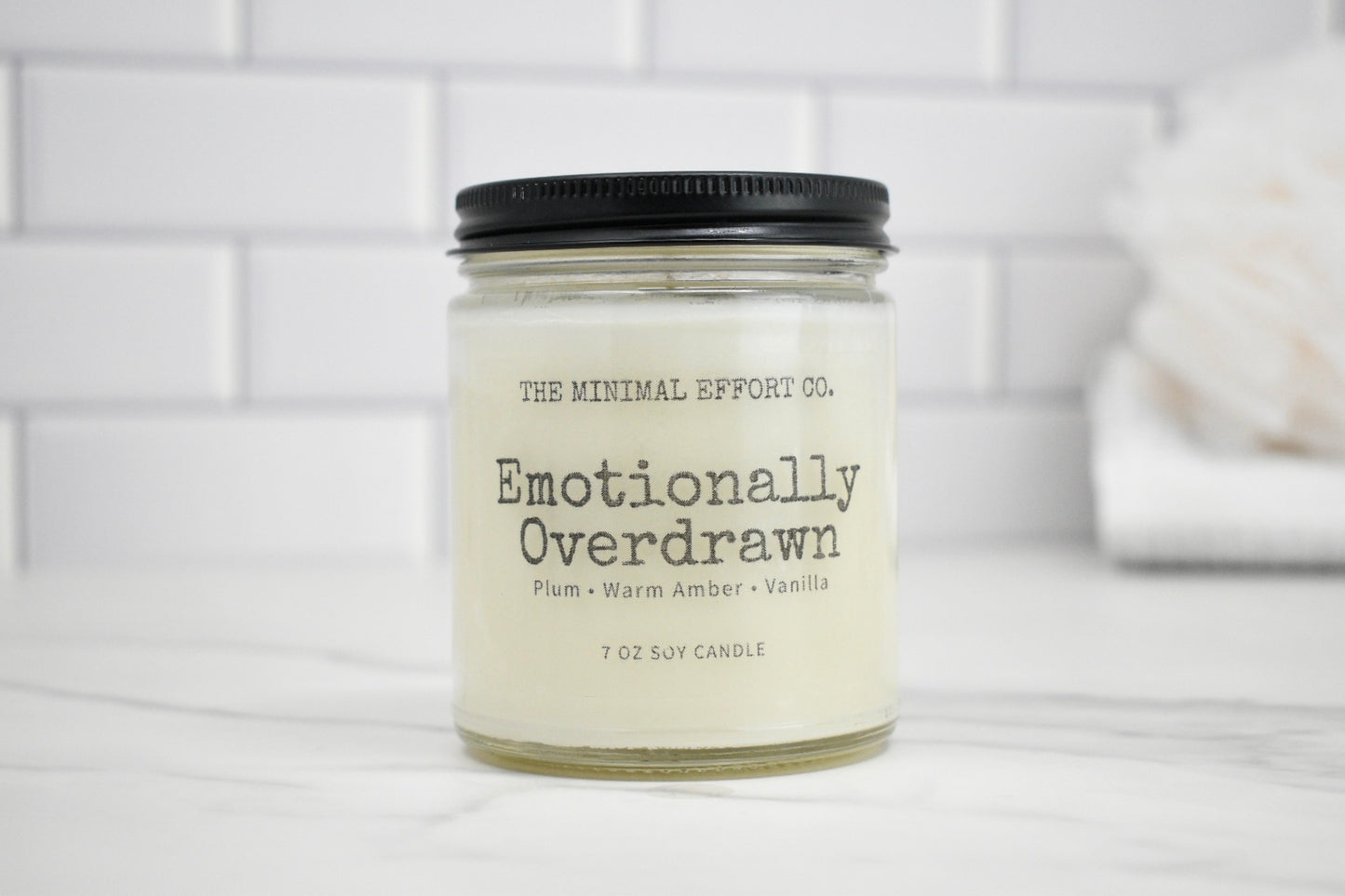 Candle jar labeled 'Emotionally Overdrawn' by The Minimal Effort Co. on a white surface with a tiled wall background.