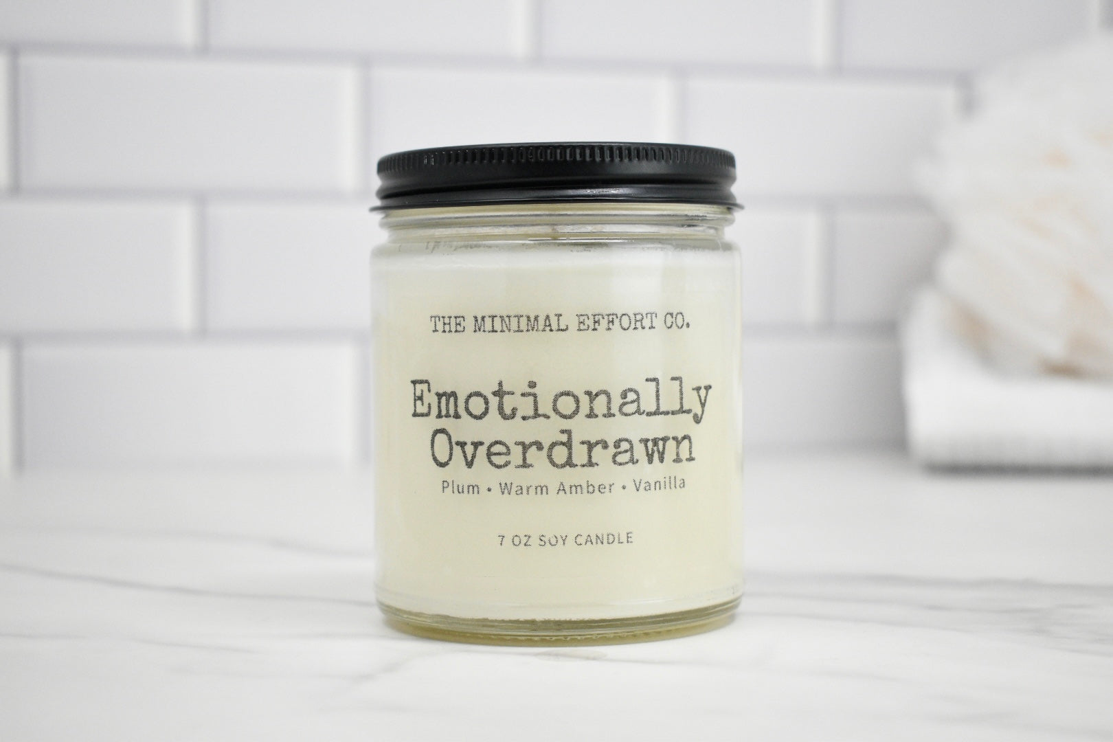 Candle jar labeled 'Emotionally Overdrawn' by The Minimal Effort Co. on a white surface with a tiled wall background.