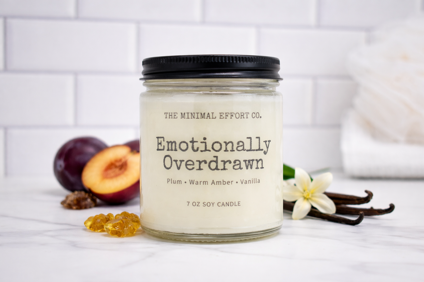 Candle labeled 'Emotionally Overdrawn' from The Minimal Effort Co. on a white surface with plums, vanilla beans, and flowers.