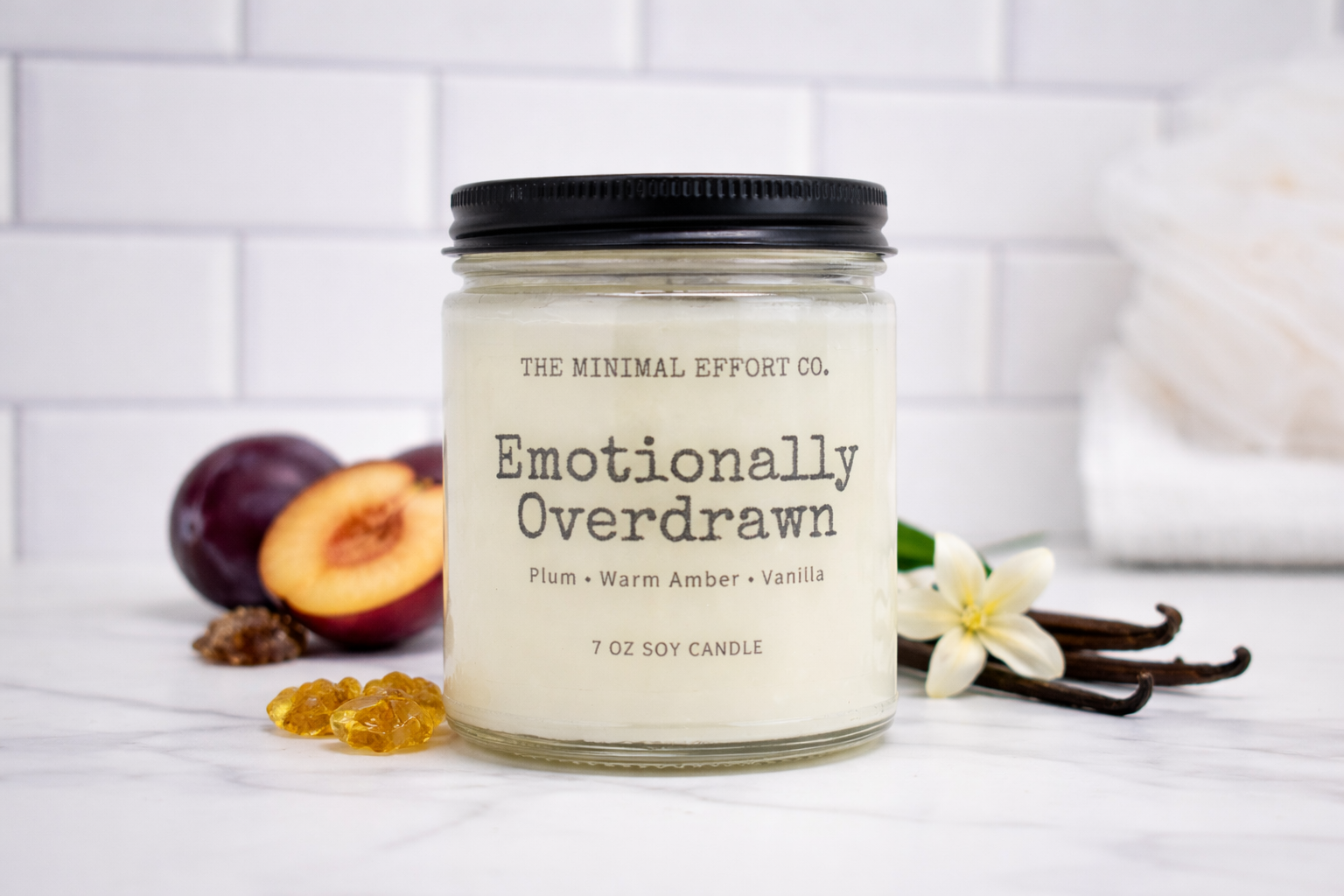 Candle labeled 'Emotionally Overdrawn' from The Minimal Effort Co. on a white surface with plums, vanilla beans, and flowers.