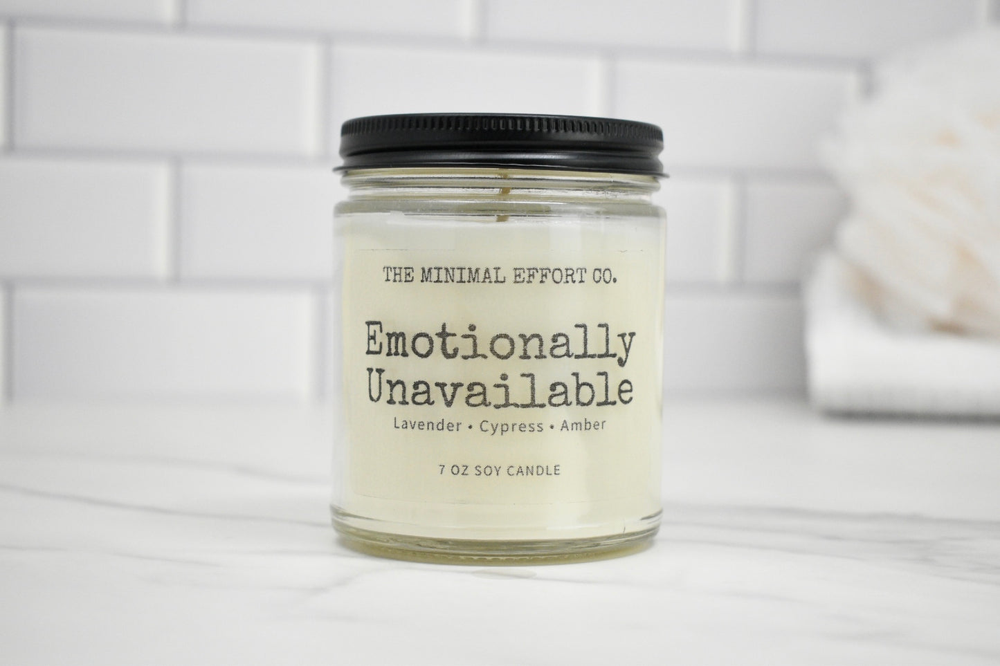 Candle jar labeled 'Emotionally Unavailable' by The Minimal Effort Co. on a white tiled background.