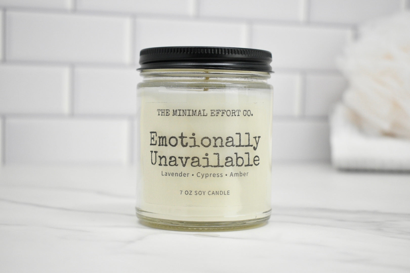 Candle jar labeled 'Emotionally Unavailable' by The Minimal Effort Co. on a white tiled background.