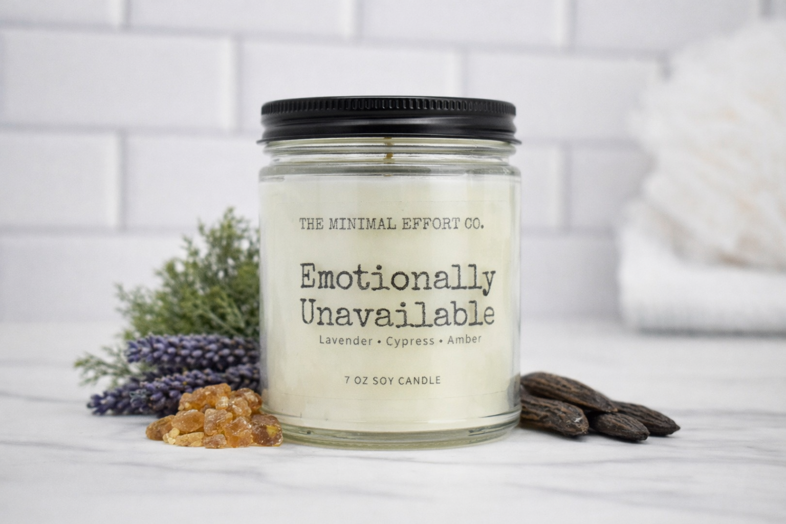 Candle jar labeled 'Emotionally Unavailable' from The Minimal Effort Co. on a white surface with lavender and amber elements.