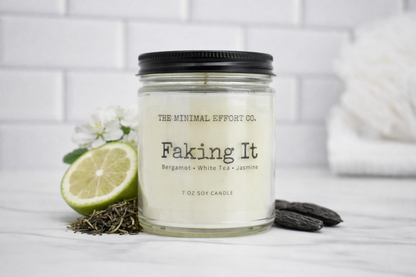 Candle labeled 'Faking It' from The Minimal Effort Co. with a lime and herbs on a white surface.