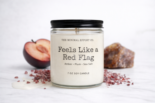 Candle labeled 'Feels Like a Red Flag' from The Minimal Effort Co. with plums and sea salt on a white background