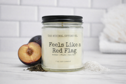 Candle labeled 'Feels Like a Red Flag' from The Minimal Effort Co. on a white surface with a blurred background.