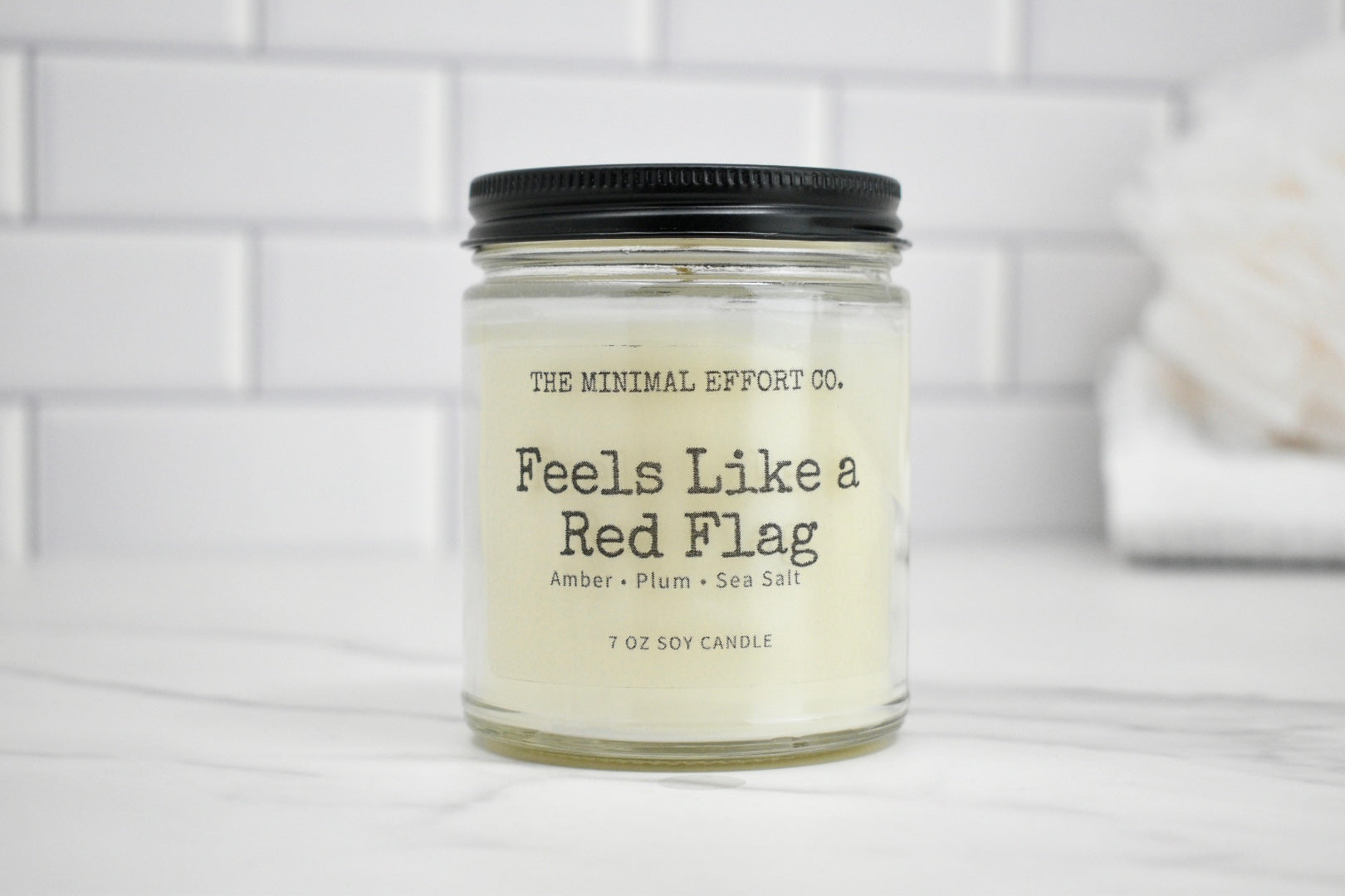 Candle jar labeled 'Feels Like a Red Flag' by The Minimal Effort Co. on a white surface with a tiled wall background.