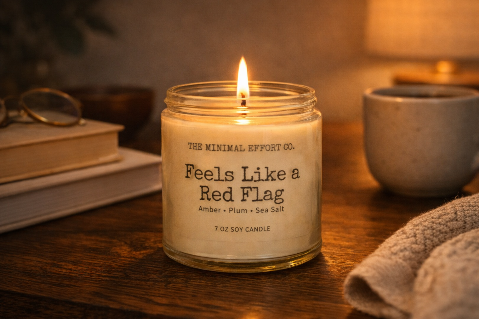 Candle labeled 'Feels Like a Red Flag' from The Minimal Effort Co. on a wooden surface with books and a lamp in the background.
