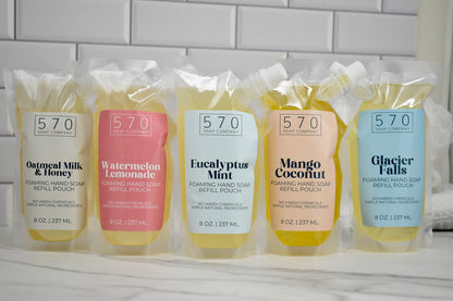Five packages of 570 Soap Company hand soap refills on a white surface with a tiled wall background.