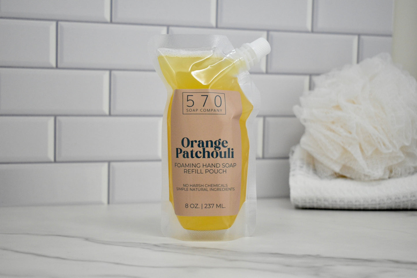 Foaming hand soap refill pouch labeled 'Orange Patchouli' on a bathroom counter with a tiled wall background.