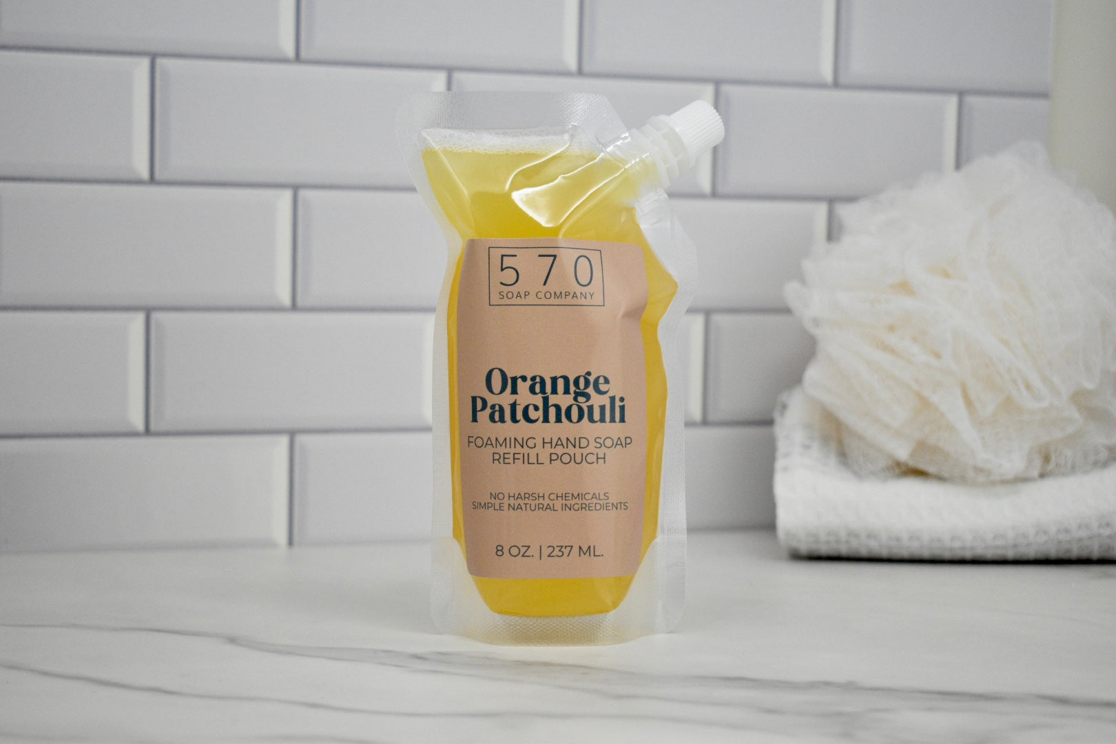 Foaming hand soap refill pouch labeled 'Orange Patchouli' on a bathroom counter with a tiled wall background.