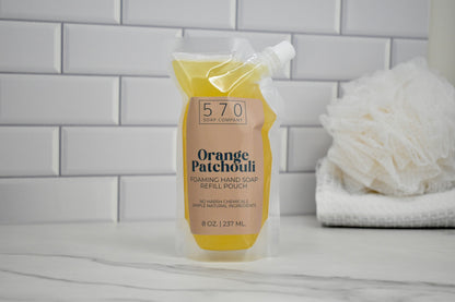 Foaming hand soap refill pouch labeled 'Orange Patchouli' on a bathroom counter with a tiled wall background.