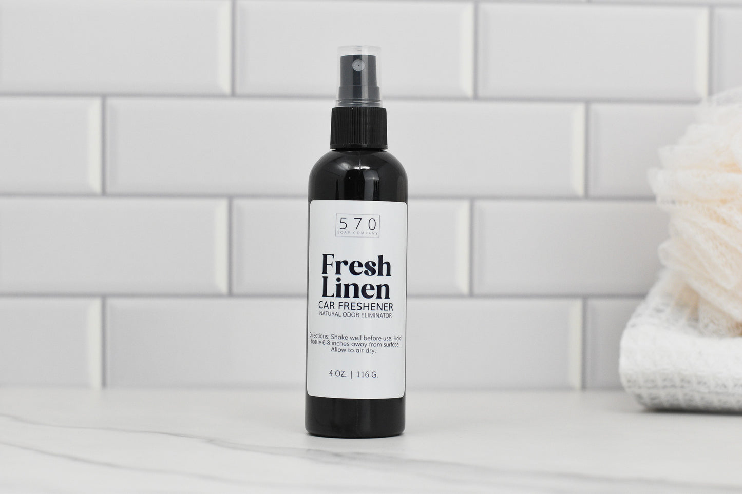 A 4 oz plastic bottle of 570 Soap Co.'s Linen & Room Spray rests on a white tiled surface, serving as an effective Fresh Linen Car Freshener. In the background, a fluffy white towel adds a cozy touch, while this natural odor eliminator fills the room with refreshing fragrances.