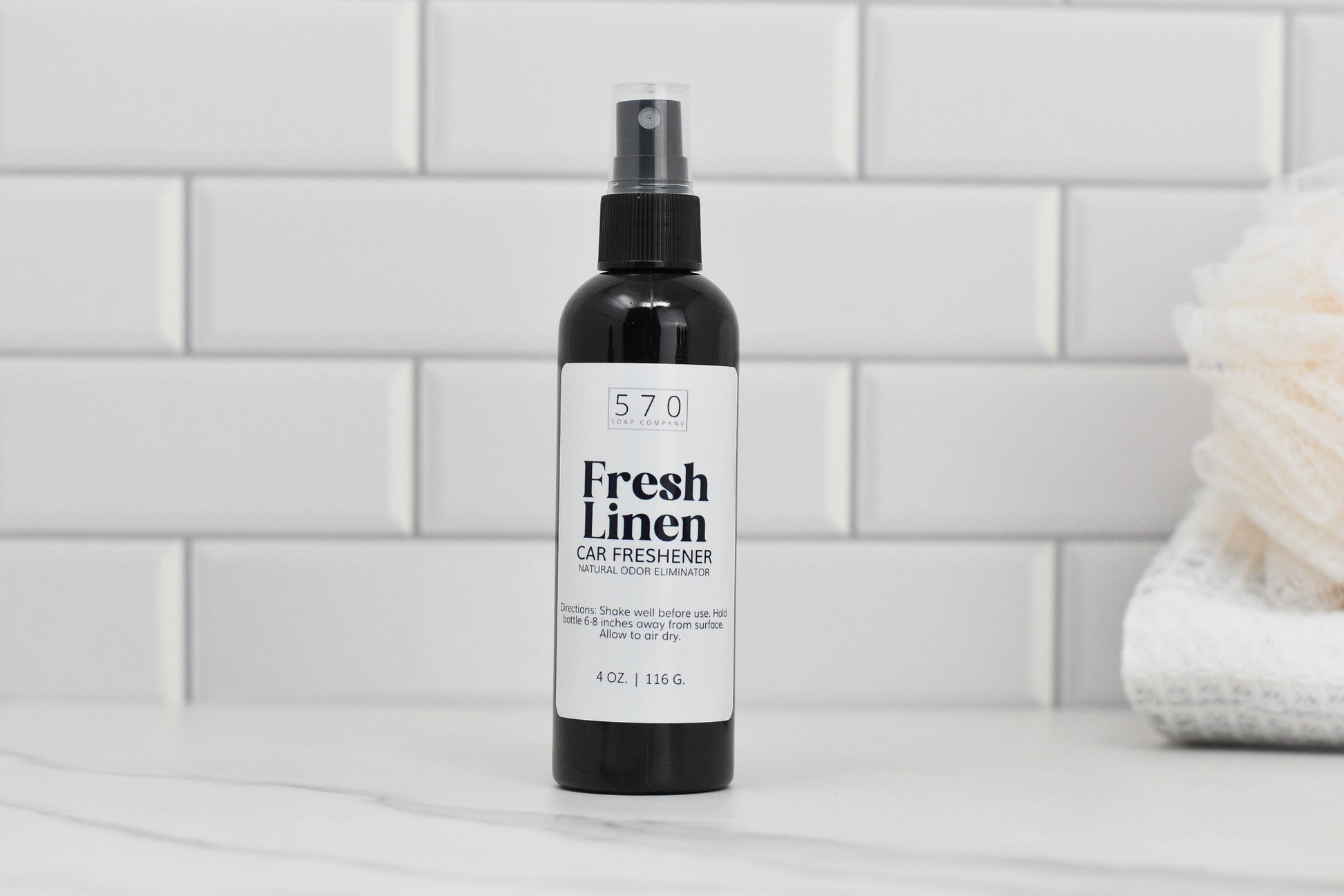 A 4 oz plastic bottle of 570 Soap Co.'s Linen & Room Spray rests on a white tiled surface, serving as an effective Fresh Linen Car Freshener. In the background, a fluffy white towel adds a cozy touch, while this natural odor eliminator fills the room with refreshing fragrances.