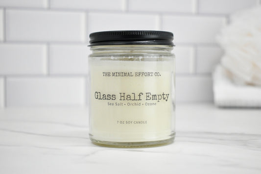 Candle jar labeled 'Glass Half Empty' from 'The Minimal Effort Co.' on a white surface with a tiled wall background.