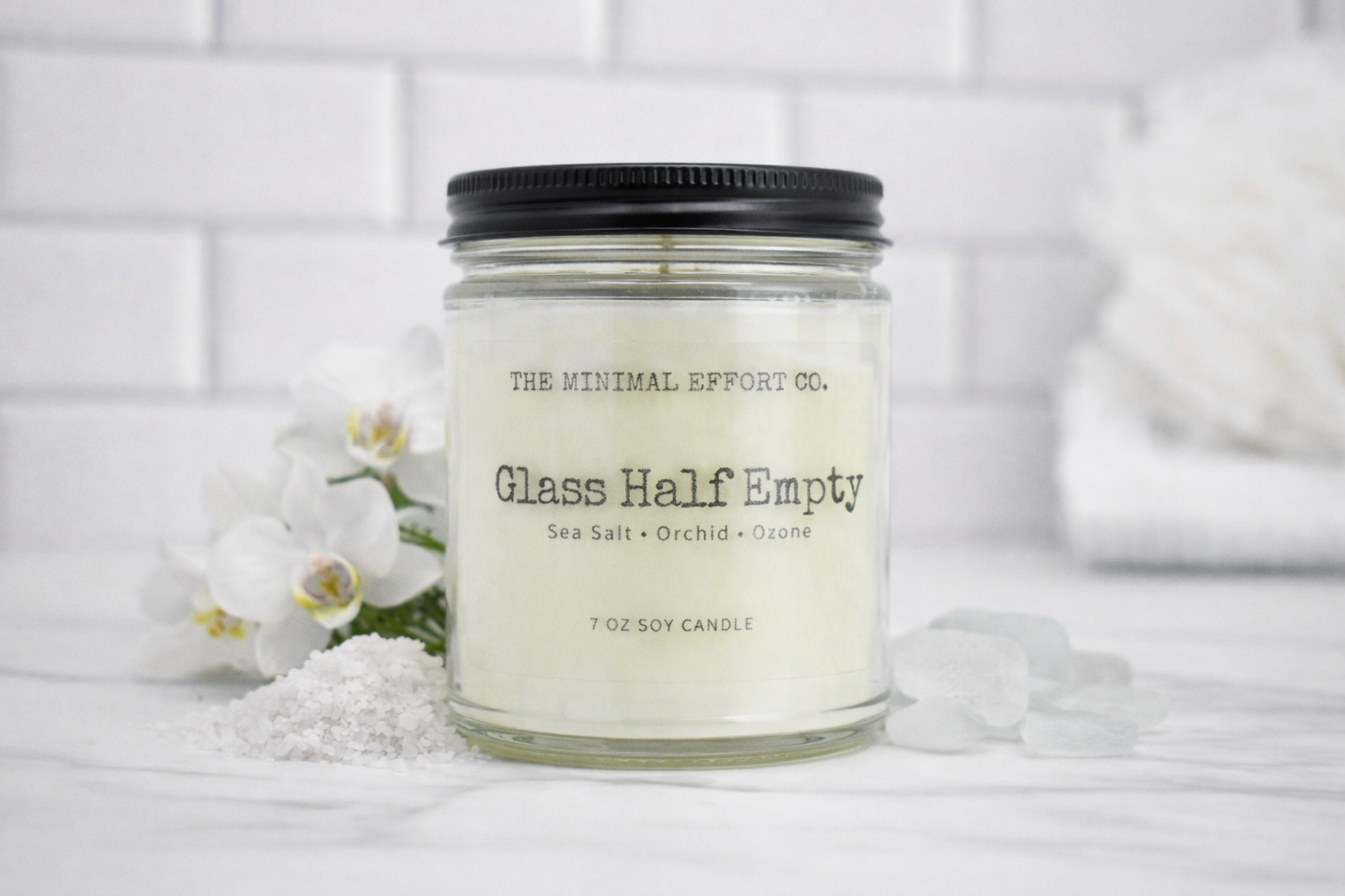 Candle jar labeled 'Glass Half Empty' from 'The Minimal Effort Co.' on a white background with flowers and rocks.
