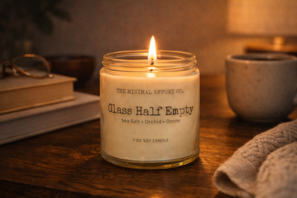 Candle labeled 'Glass Half Empty' from 'The Minimal Effort Co.' on a wooden surface with books and a lamp in the background.