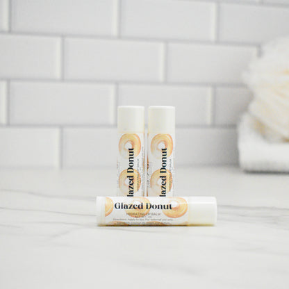 Three lip balms labeled 'Glazed Donut' on a bathroom counter with a tiled wall background.