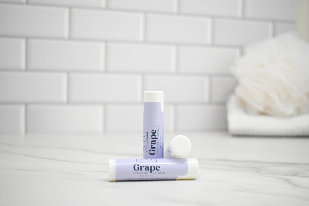 Two lip balm tubes labeled 'Grape' on a marble surface with a tiled wall background.