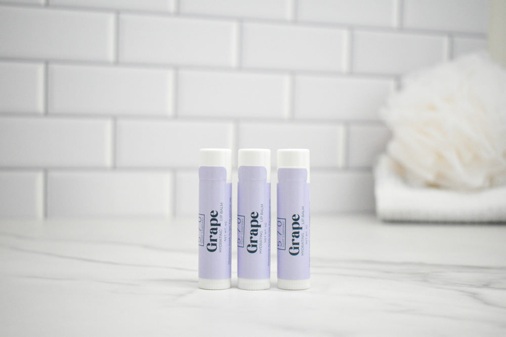 Three lip balm tubes labeled 'Grape' on a white surface with a tiled wall background.