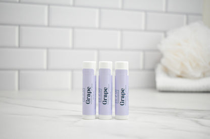 Three lip balm tubes labeled 'Grape' on a white surface with a tiled wall background.