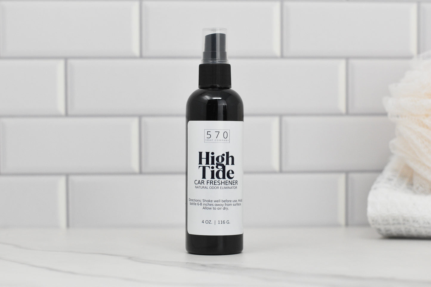 A bottle of Linen & Room Spray in a 4 oz plastic container from 570 Soap Co., renowned for its natural odor eliminator properties, is placed on a marble surface. The label features a simple design, set against a backdrop of white subway tiles and complemented by a folded white towel.