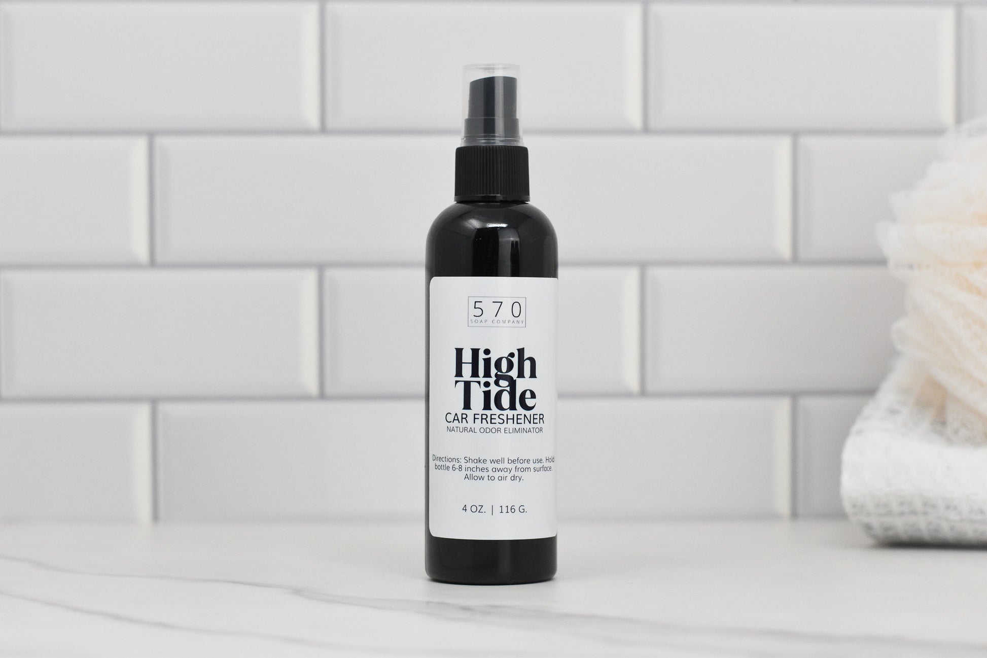 A bottle of Linen & Room Spray in a 4 oz plastic container from 570 Soap Co., renowned for its natural odor eliminator properties, is placed on a marble surface. The label features a simple design, set against a backdrop of white subway tiles and complemented by a folded white towel.