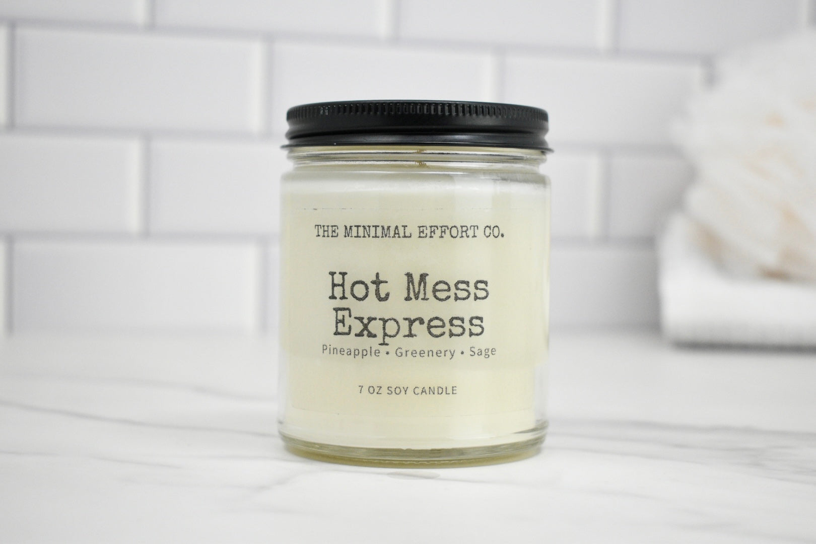 Candle labeled 'Hot Mess Express' from The Minimal Effort Co. on a white surface with a tiled wall background.