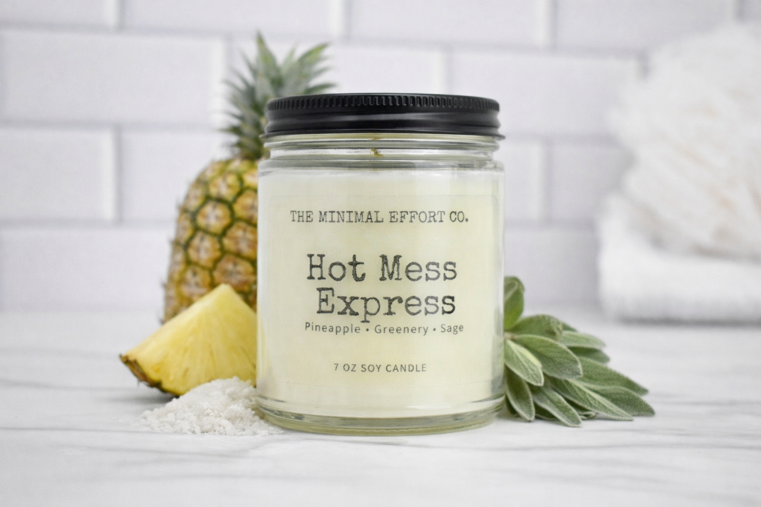 Candle labeled 'Hot Mess Express' from 'The Minimal Effort Co.' with a pineapple and greenery on a white background
