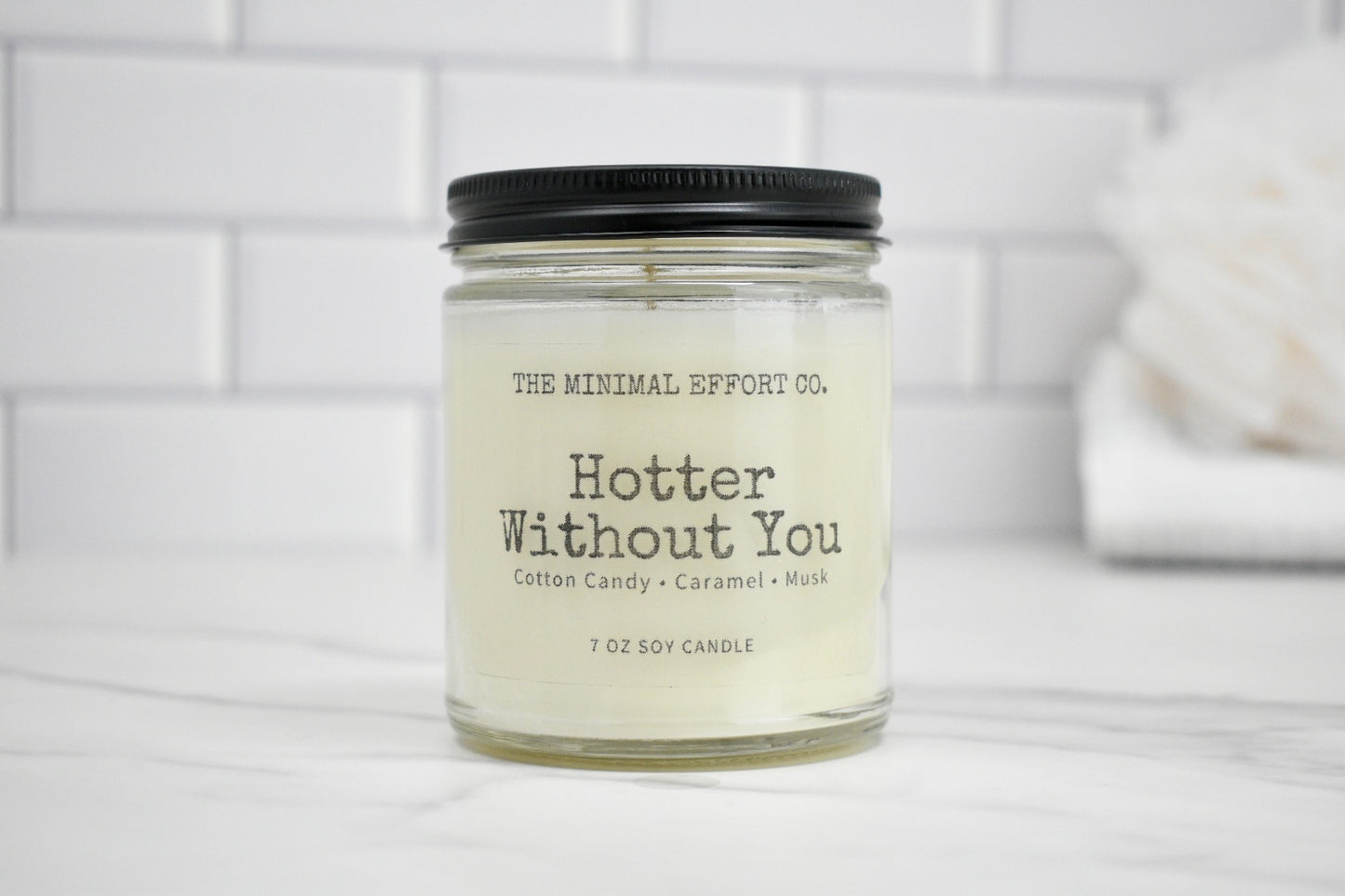 Candle jar labeled 'Hotter Without You' by The Minimal Effort Co. on a white surface with a tiled wall background.