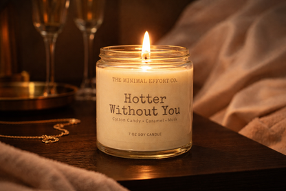 Candle labeled 'Hotter Without You' from The Minimal Effort Co. on a wooden surface with soft lighting.