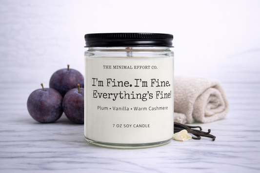 Candle jar with text 'I'm Fine, I'm Fine Everything's Fine' surrounded by plums and vanilla beans on a light background