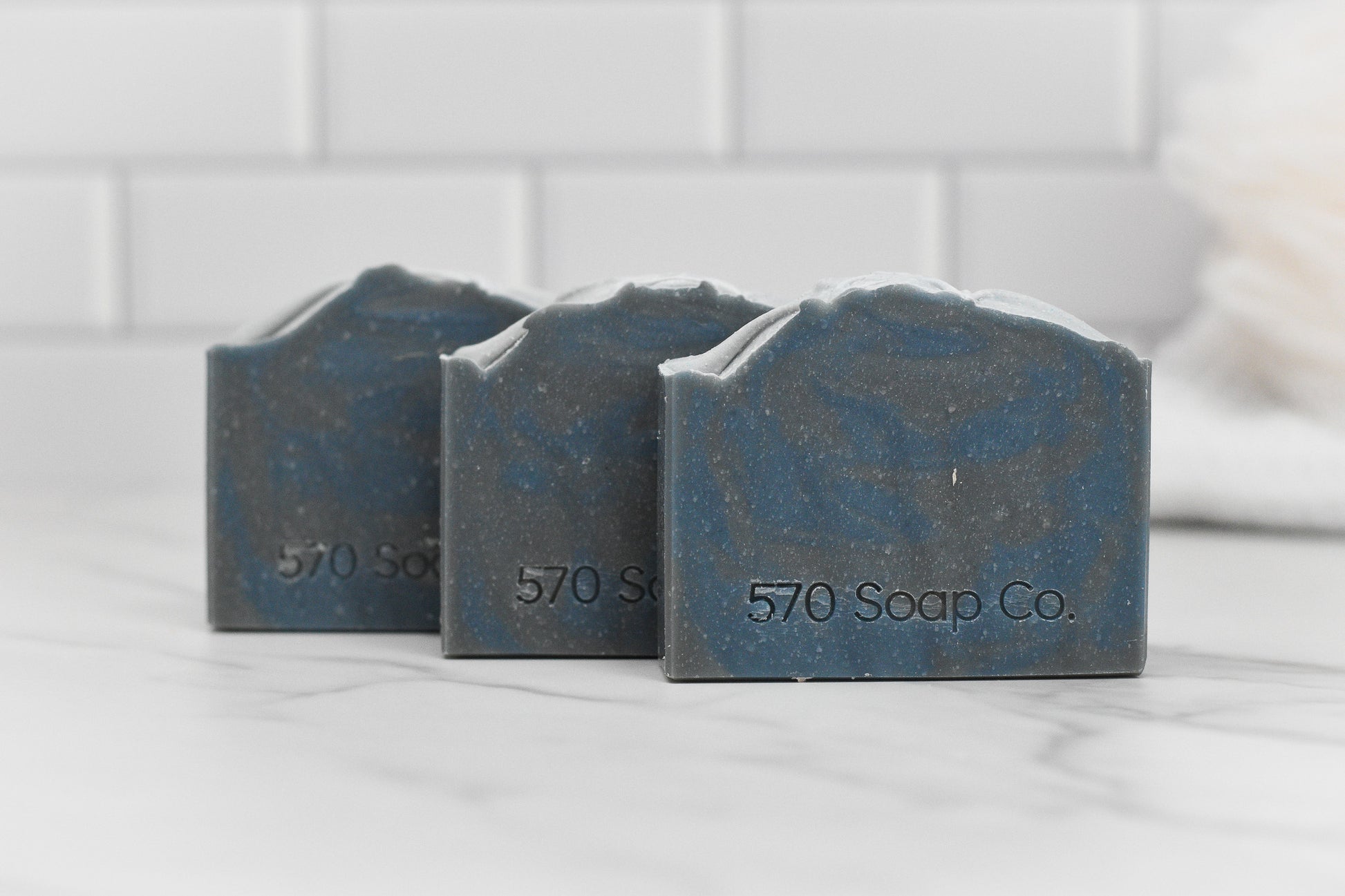 Three bars of "Date Night Bar Soap: The Ultimate Man Soap" from 570 Soap Co. are arranged on a marble surface with a white subway tile background, exuding a masculine allure that promises a captivating fragrance journey.