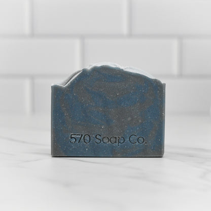 A bar of "Date Night Bar Soap: The Ultimate Man Soap" by 570 Soap Co., featuring a marbled blue and gray design, sits on a white marble surface, exuding masculine allure. The name 570 Soap Co. is imprinted on the front. Classic white subway tiles in the background create a captivating base for any fragrance journey.