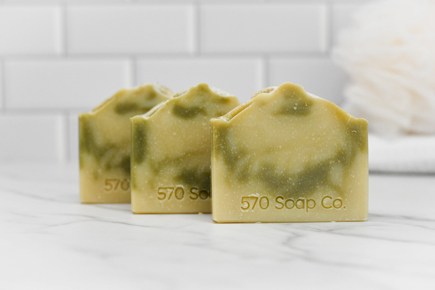 Arranged in a row on a white marble surface, three marbled green and cream bars of the Patchouli Essential Oil Bar Soap from 570 Soap Co. are engraved with the brand name. The earthy elegance of Patchouli Essential Oil infuses the scene, set against a background of white subway tiles and a blurred bath sponge.