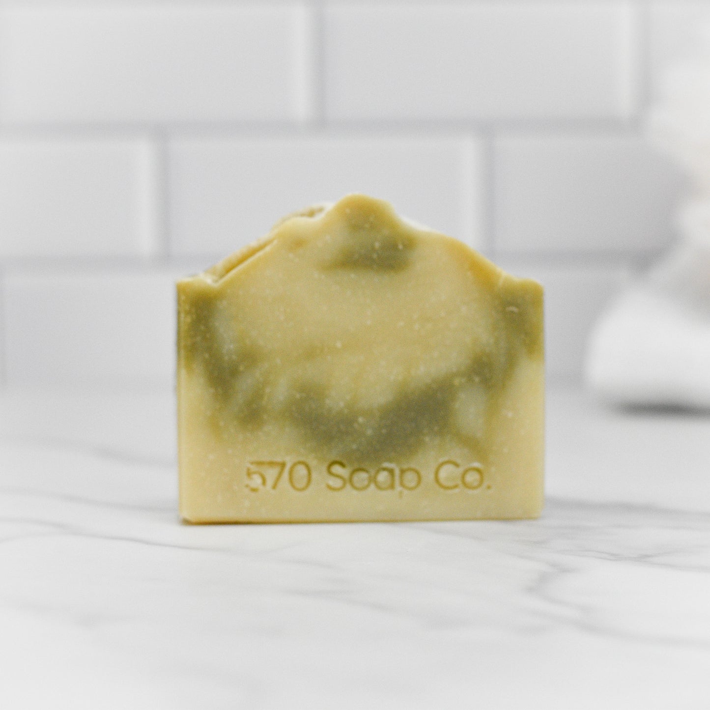 A bar of Patchouli Essential Oil Bar Soap - All Natural from 570 Soap Co. showcases an earthy elegance with its marbled green and beige design, and is captured resting on a white marble surface against a white tiled background.