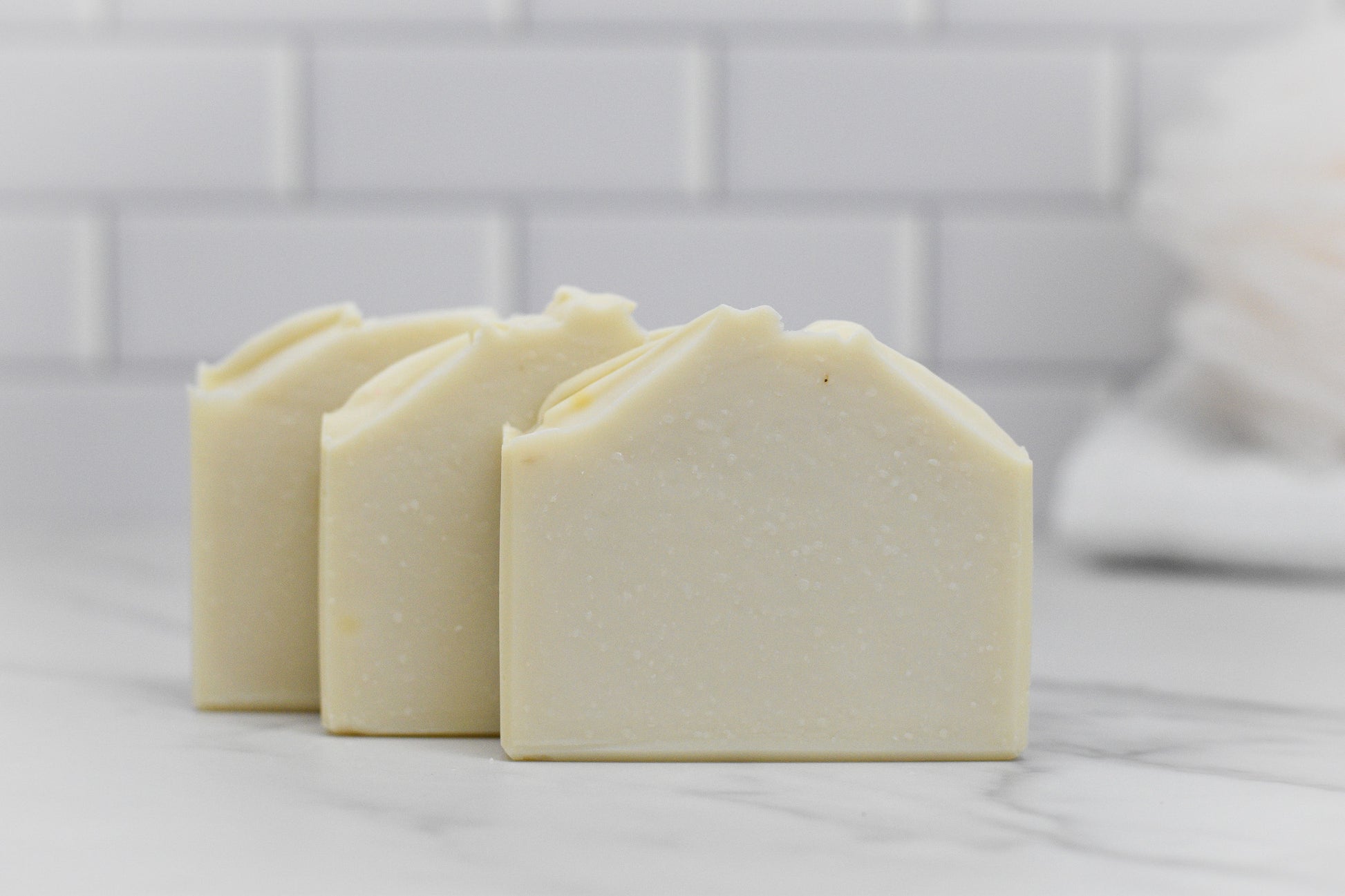 Three bars of 570 Soap Co.'s Jewelweed Infused Bar Soap - All Natural are neatly lined up on a marble surface. The background features a blurred white tile wall, adding a clean and minimalist touch to the scene.