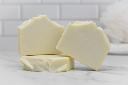 Three Jewelweed Infused Bar Soaps from 570 Soap Co. are arranged with two standing upright and leaning against each other, while a third lays flat in front. The beige soap bars, infused with jewelweed and enriched with coconut oil, are set against a backdrop of a white tiled wall and blurred bathroom items, creating a clean, minimalistic setting.