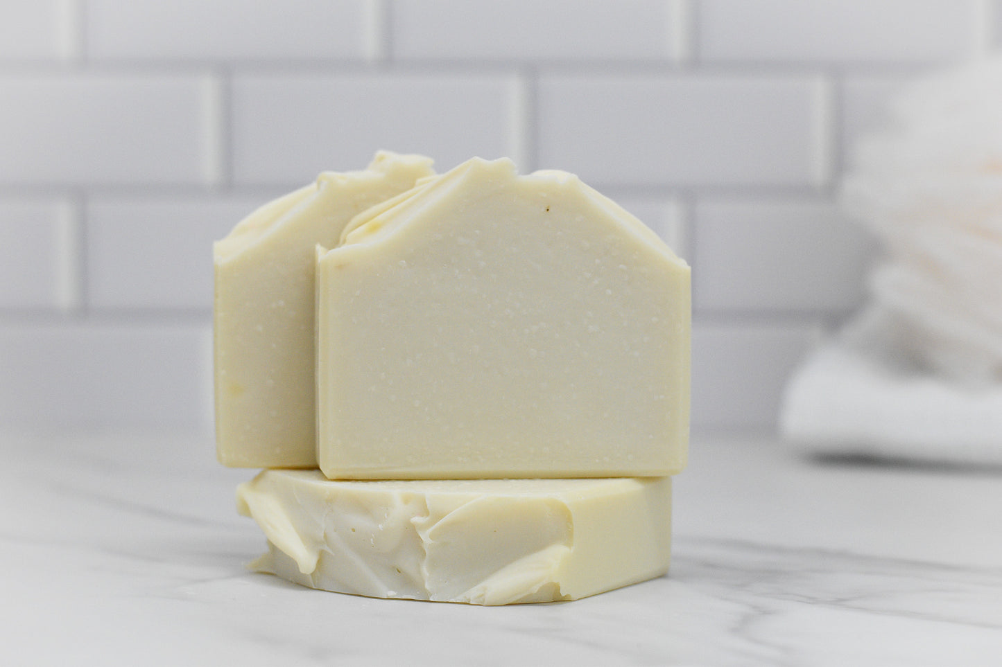 Two bars of the Jewelweed Infused Bar Soap - All Natural by 570 Soap Co., made with coconut oil, are stacked against white subway tiles, complemented by a soft towel in the background. The soap showcases a smooth texture and charming natural imperfections along its edges.