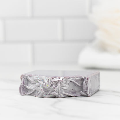The Juicy Grape Delight Bar Soap by 570 Soap Co., featuring a swirled, marbled design in shades of gray, white, and purple and enriched with kaolin clay and olive oil, is elegantly displayed on a white marble surface. In the background are white subway tiles with blurred bathroom accessories.