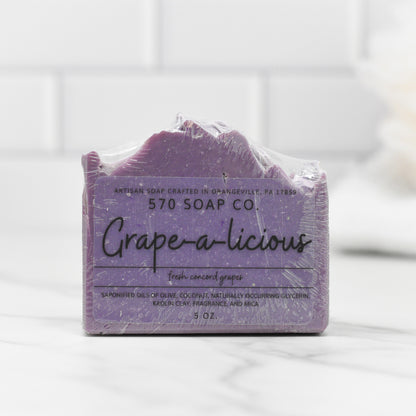 A Juicy Grape Delight bar of soap from 570 Soap Co., wrapped in clear plastic. The label reveals it as an artisan creation with a fresh concord grape scent, featuring saponified oils, glycerin, kaolin clay, and fragrance.