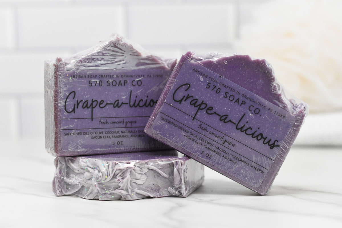 Two bars of 570 Soap Co.'s Juicy Grape Delight Bar Soap adorn a white tiled backdrop, showcasing their purple tones and rugged charm. Infused with olive oil, these grape-scented treasures add a luxurious touch to any setting. The packaging lists ingredients and weight, emphasizing the soap's artisanal quality.