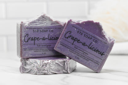 Two bars of 570 Soap Co.'s Juicy Grape Delight Bar Soap adorn a white tiled backdrop, showcasing their purple tones and rugged charm. Infused with olive oil, these grape-scented treasures add a luxurious touch to any setting. The packaging lists ingredients and weight, emphasizing the soap's artisanal quality.