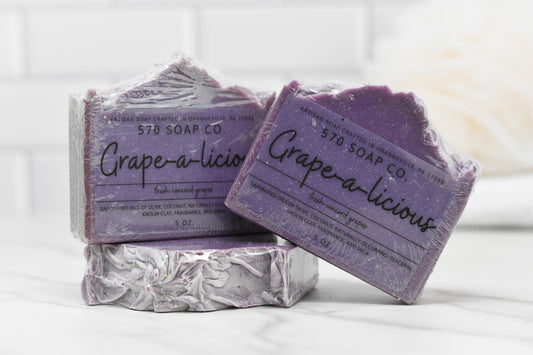 Two bars of 570 Soap Co.'s Juicy Grape Delight Bar Soap adorn a white tiled backdrop, showcasing their purple tones and rugged charm. Infused with olive oil, these grape-scented treasures add a luxurious touch to any setting. The packaging lists ingredients and weight, emphasizing the soap's artisanal quality.