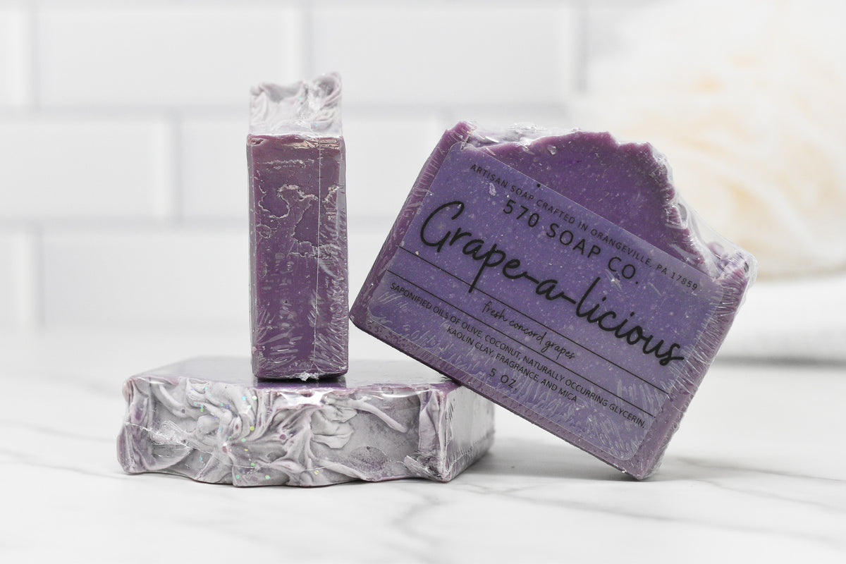 Three bars of Juicy Grape Delight bar soap are arranged on a white marble surface. One stands upright, one lies flat, and the third is angled. Labeled with the brand name 570 Soap Co., these soaps feature kaolin clay and nourishing olive oil, with ingredients and company details printed on the packaging.