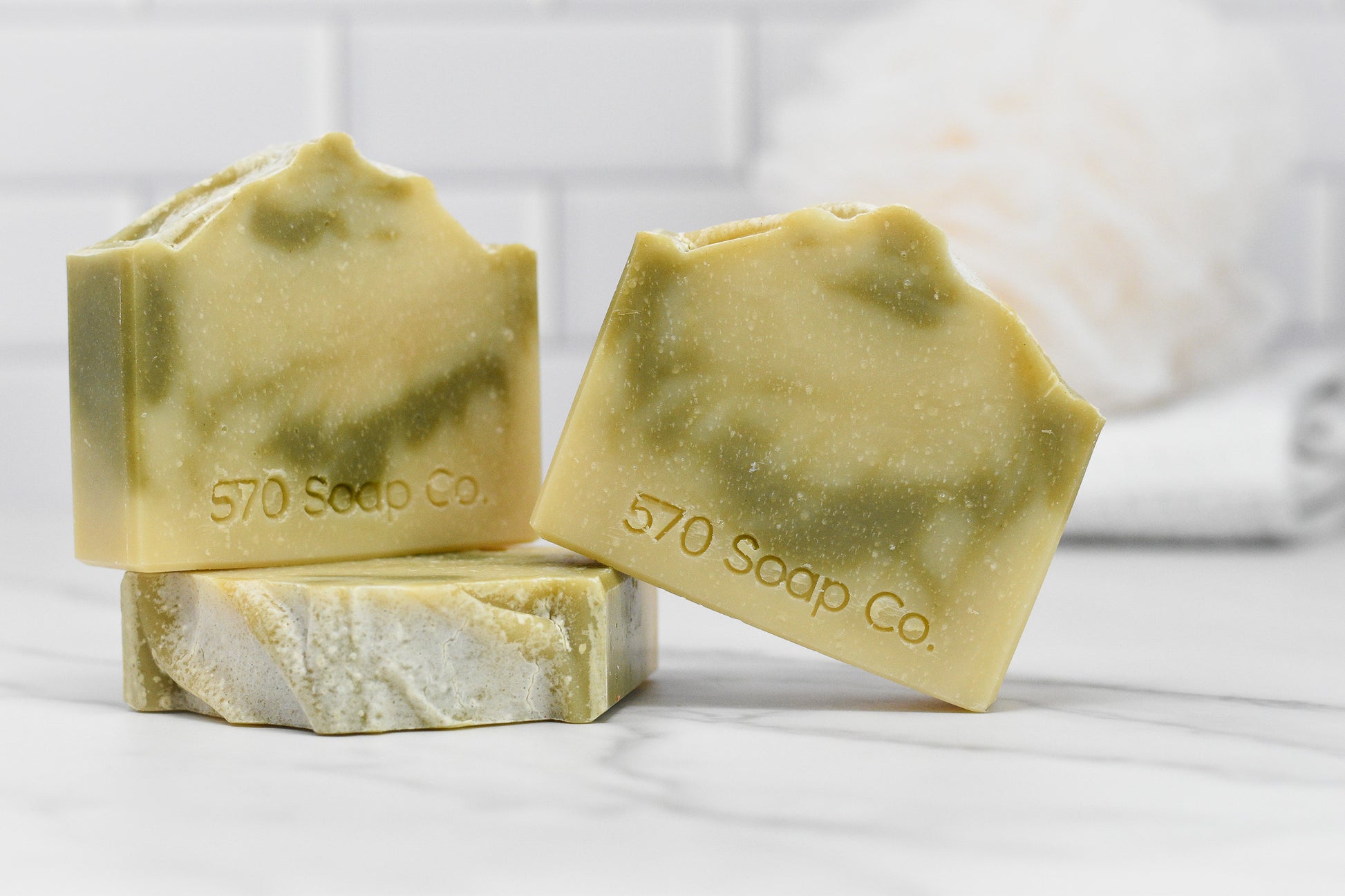 Three Patchouli Essential Oil Bar Soaps from 570 Soap Co. are displayed on a marble surface, radiating an earthy elegance. Two bars are positioned upright while another rests flat. In the background, a blurred white loofah is visible against a tiled wall. The soaps feature a marbled texture in earthy tones and are infused with all-natural essential oils.