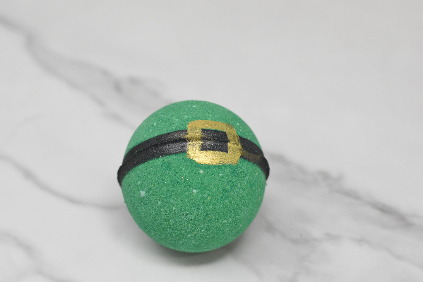 The Enchanting Elf Bath Bomb: Round by 570 Soap Co., resembling a leprechaun hat in green, is infused with skin-nourishing ingredients and whimsical charm, featuring a black band and a gold buckle as it rests on a white marble surface.